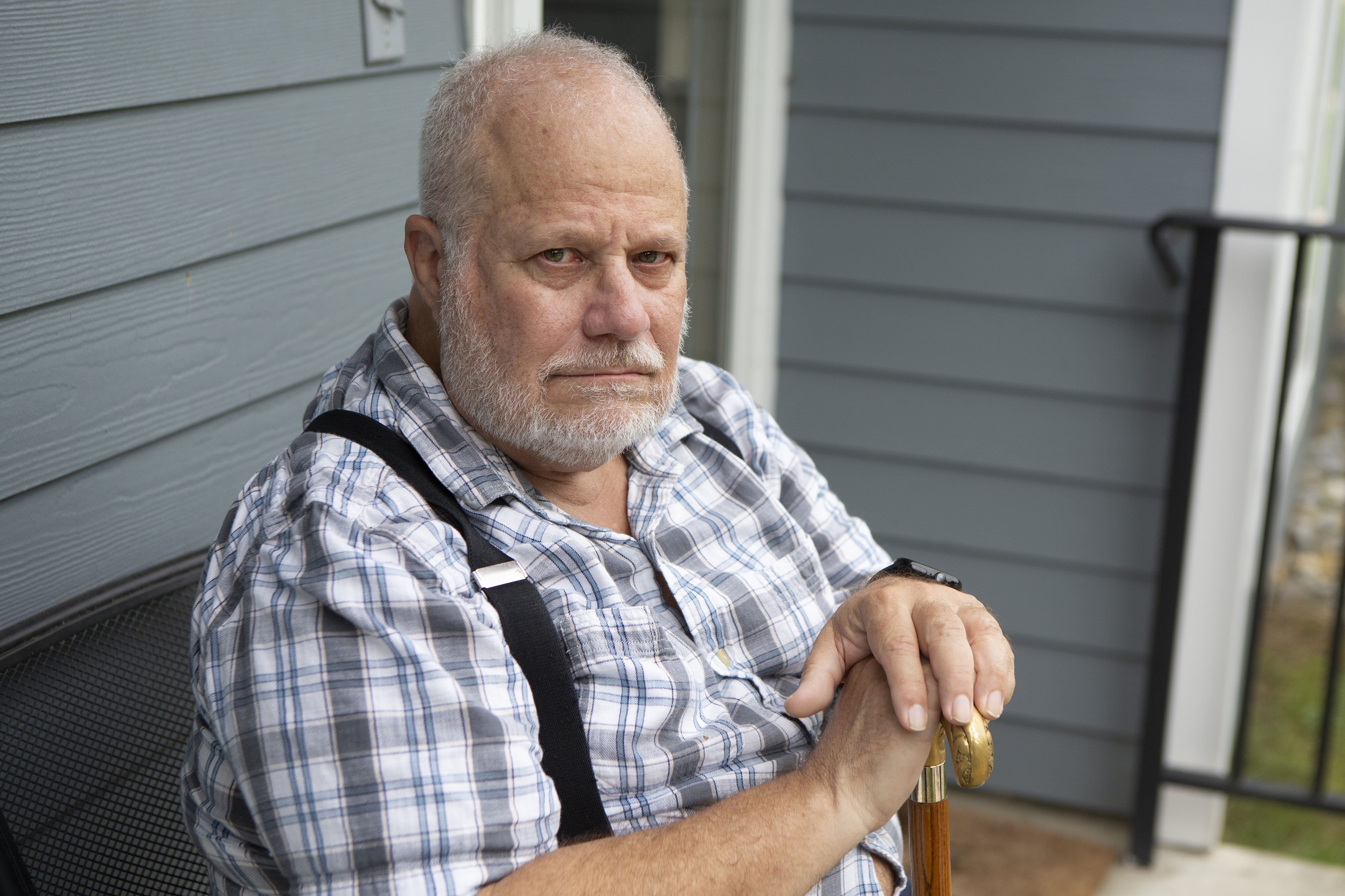 An older man wearing a plaid shirt with suspenders sits on his front porch with his hands resting on his cane.