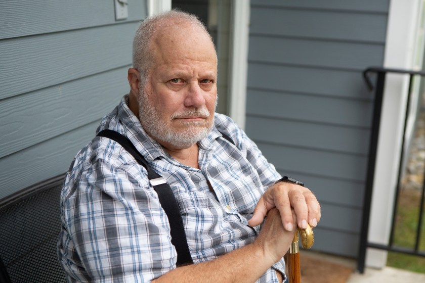 An older man wearing a plaid shirt with suspenders sits on his front porch with his hands resting on his cane.