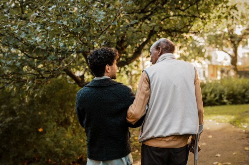 A younger man walks with an elderly man through a leafy street.