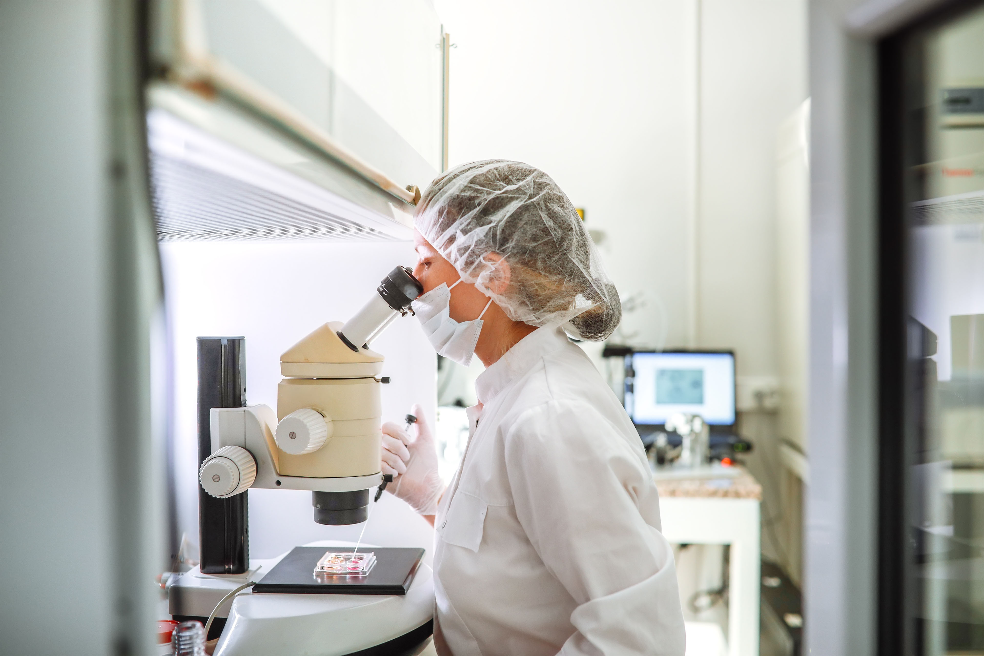 A photo of an embryologist in an IVF laboratory using a microscope while working with a pipette and an embryo in a petri dish.
