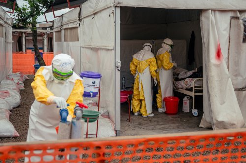 Three people in head-to-toe personal protective equipment are in the frame of this photo. One is in the foreground outside a tent accessing a tap while the other two are inside the tent, facing a person lying on a cot.