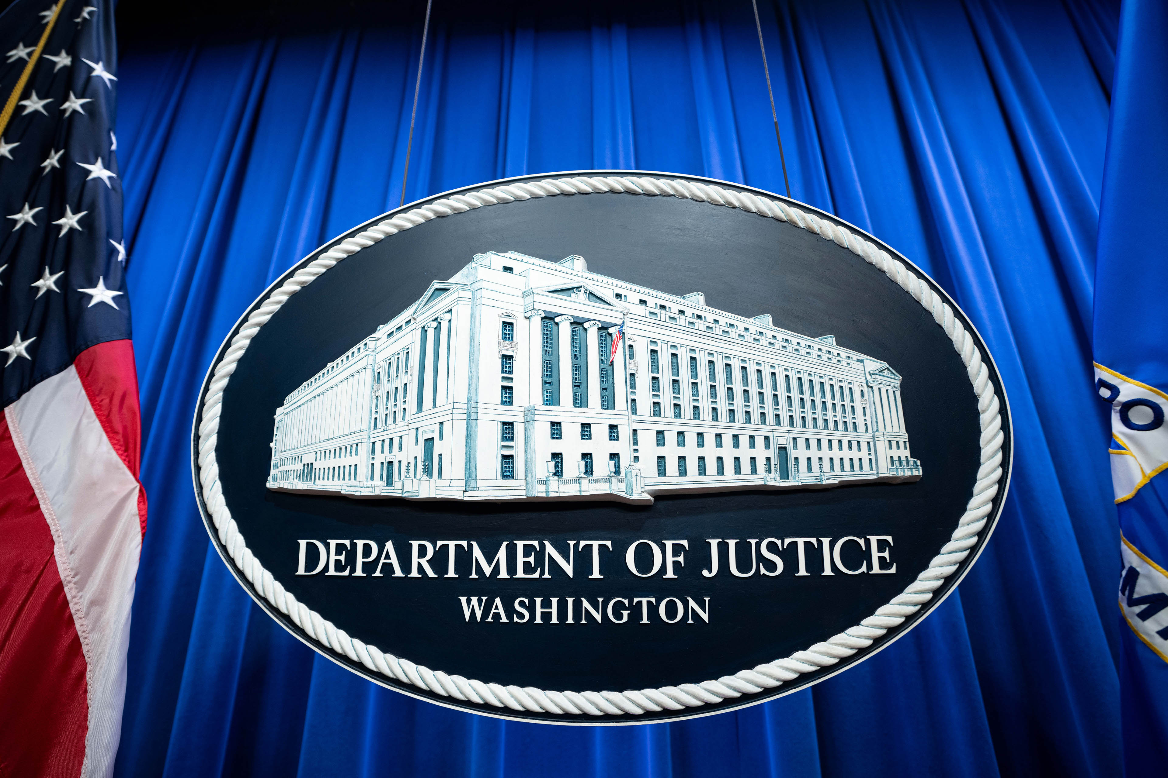 Department of Justice signage hangs in front of a blue curtain.