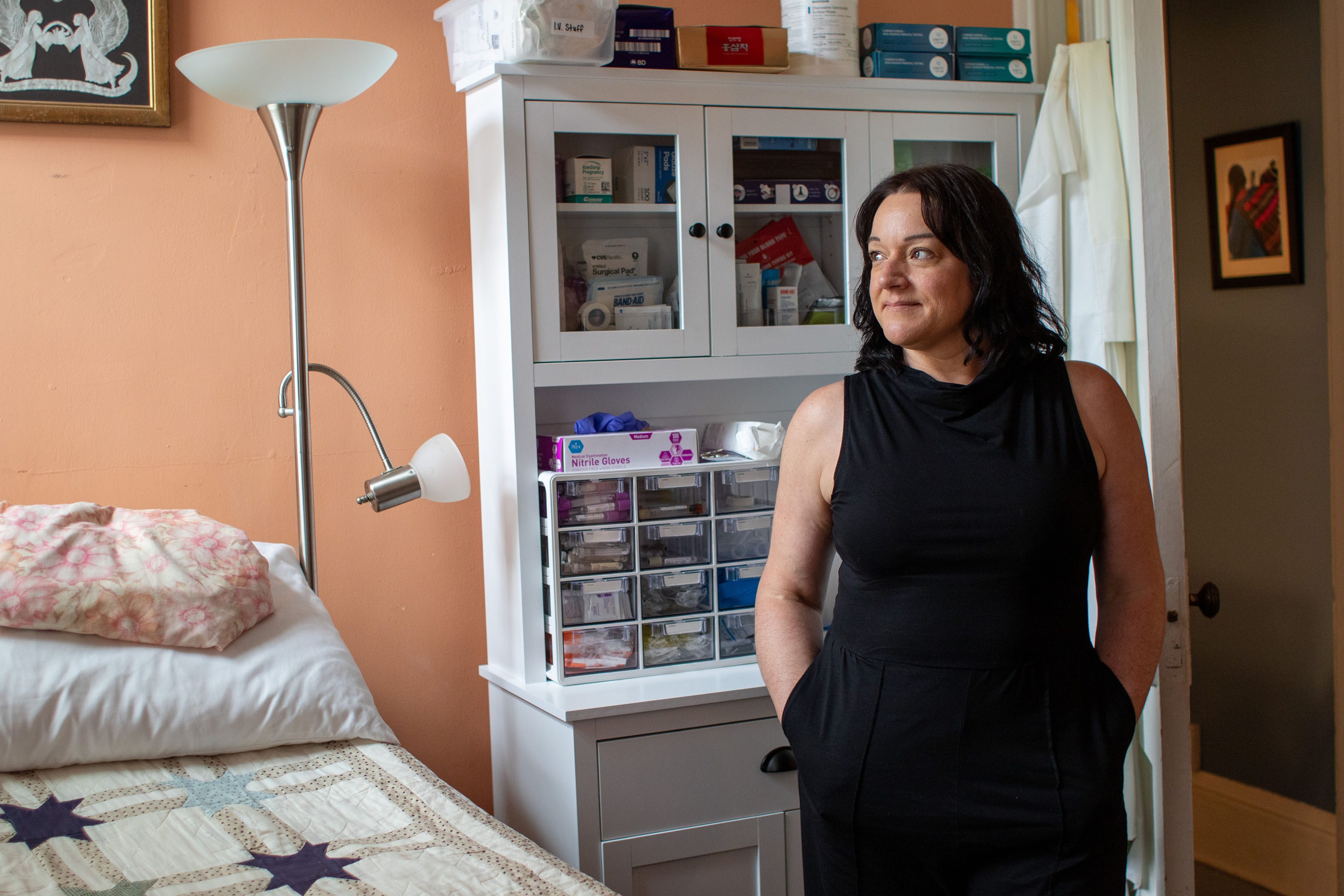 A woman stands beside a bed with a quilt looks away from the camera with a subtle smile.