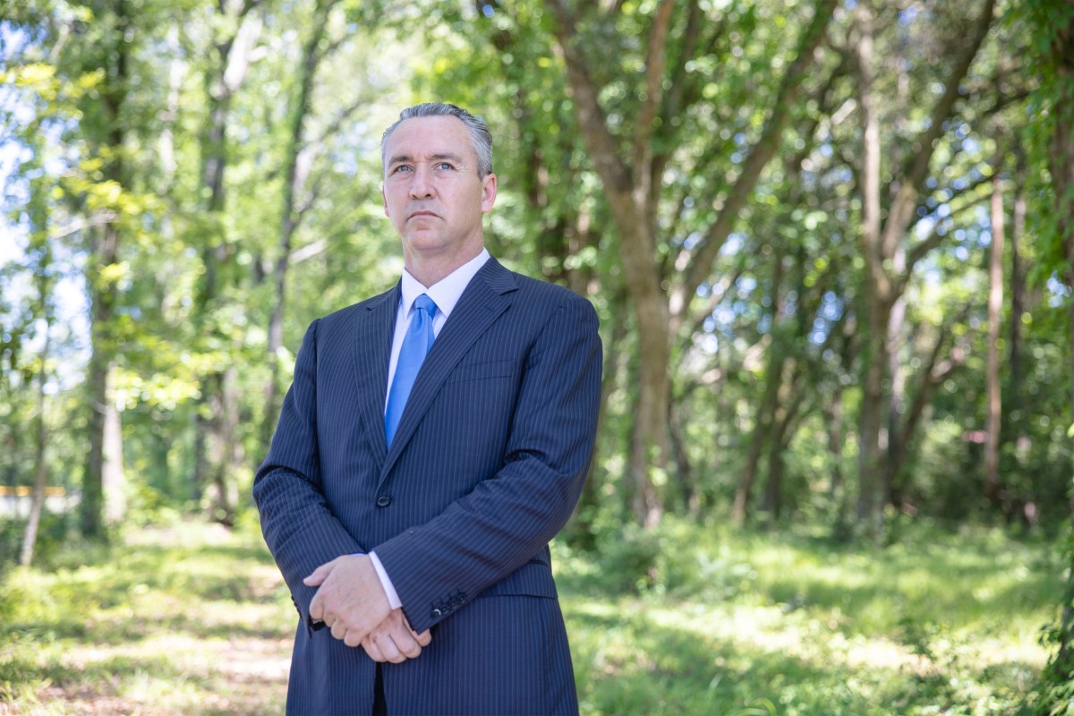 A man in a suit stands for a portrait outside. A woodsy background is behind him.