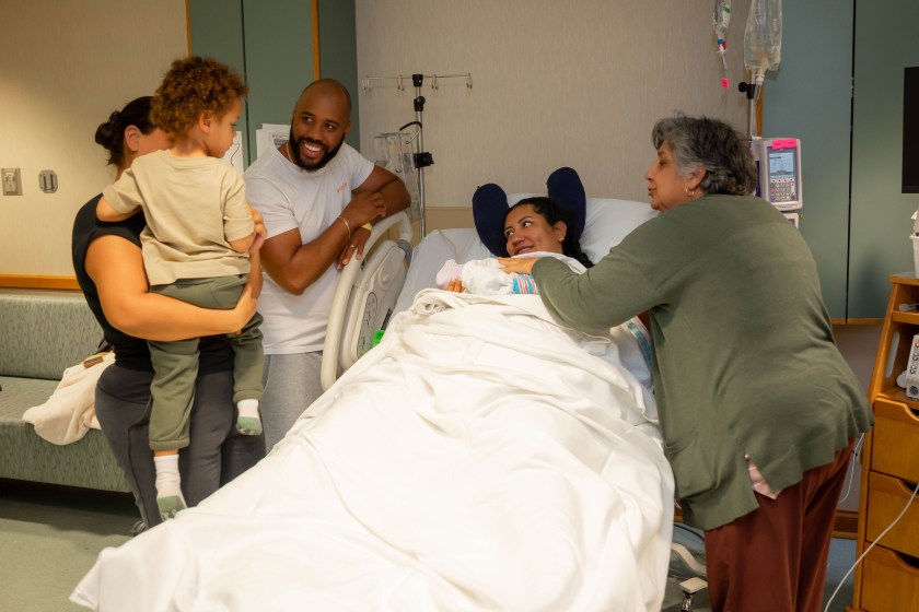 A woman who just gave birth holds her infant while lying in a hospital bed. Three adult family members and one child stand around her.