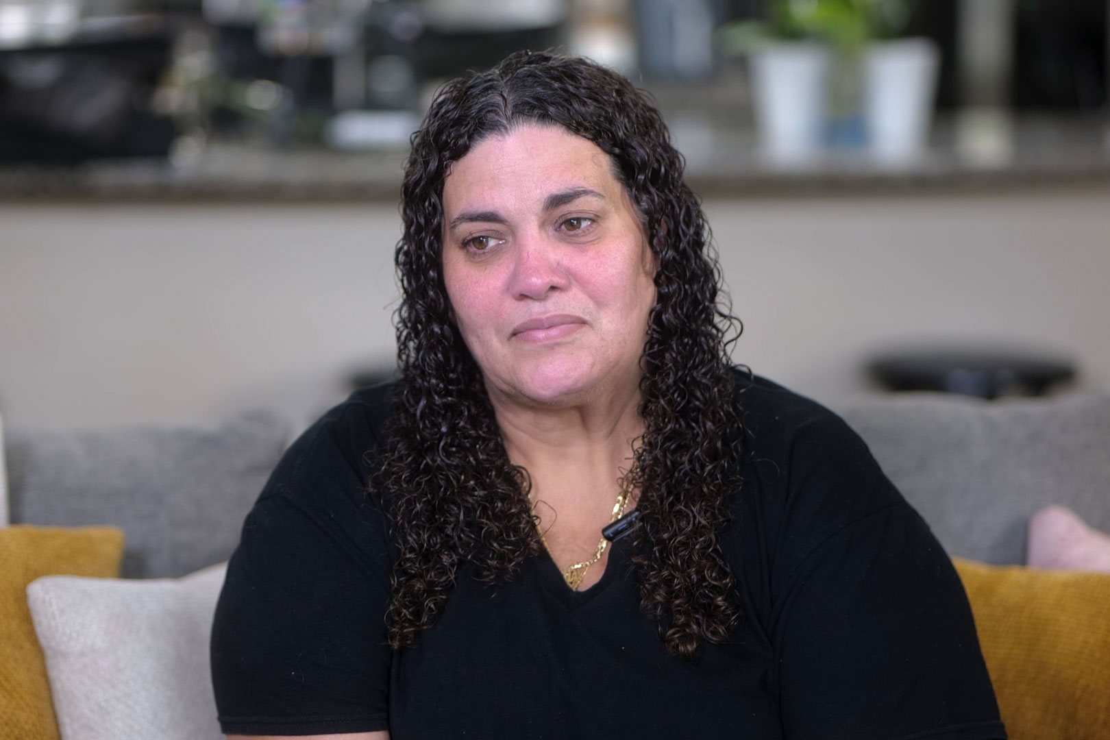 A Latina woman sits for a television interview in her home. She is tearing up.