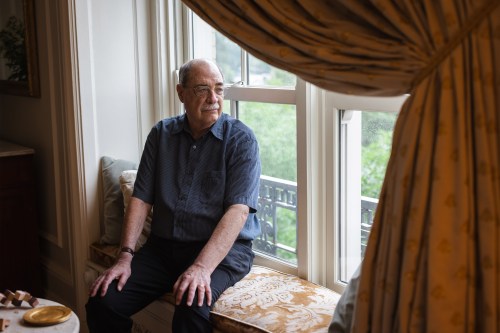 A photo of an older man sitting in a window nook. The window's curtain is drawn.