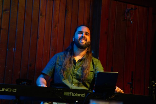 A man with a short beard and long brown hair in a button down green shirt smiles as he plays a keyboard in a music venue.