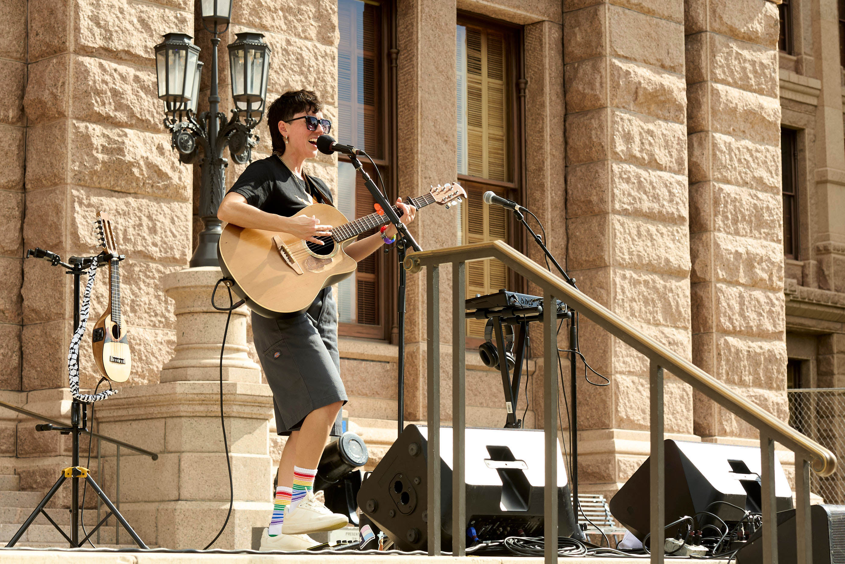 A person in a black T-shirt plays a guitar on the steps of a state building on a sunny day.