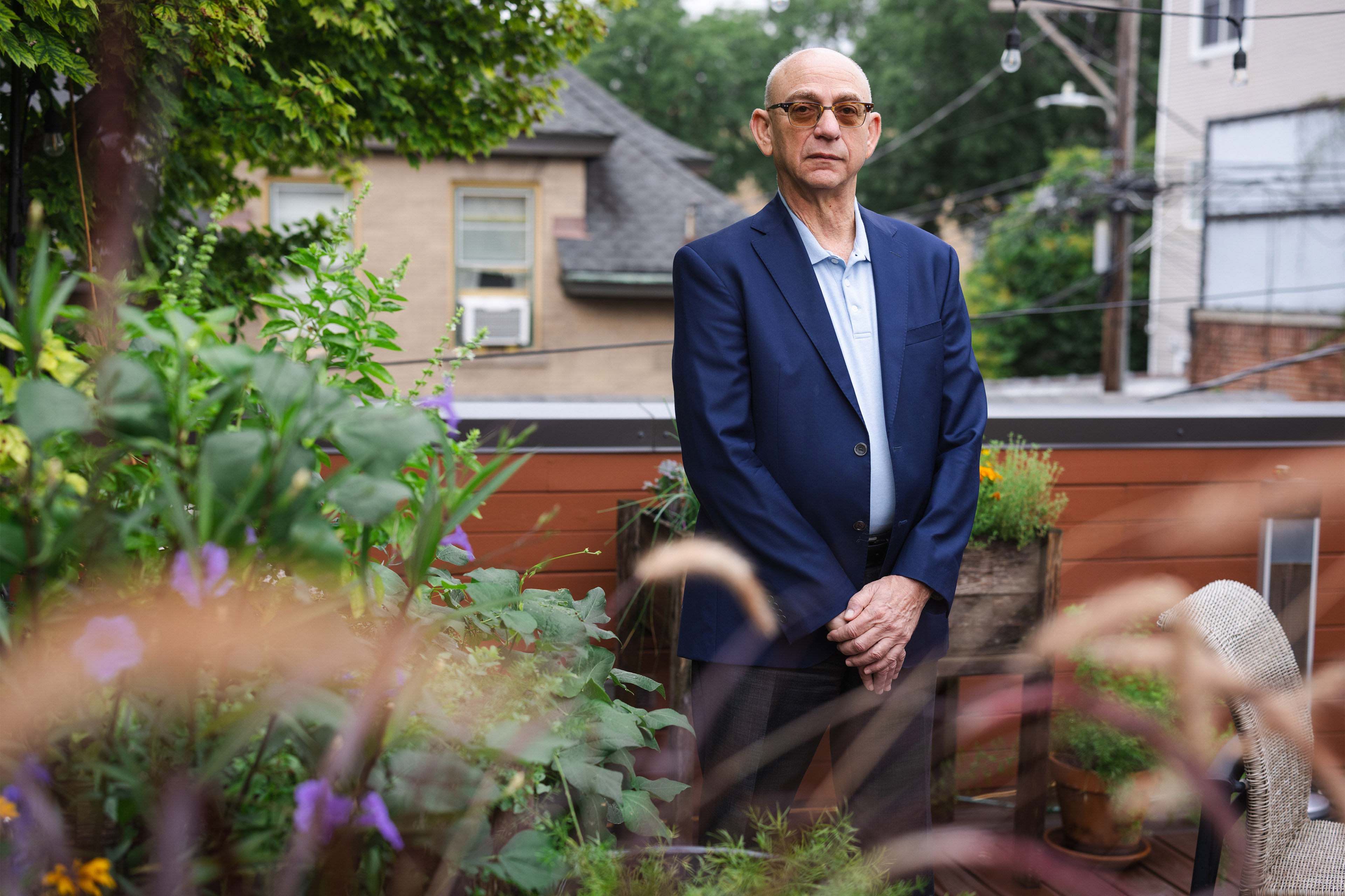A man stands in a garden area outside of his home. Foliage is seen blurred in the foreground.