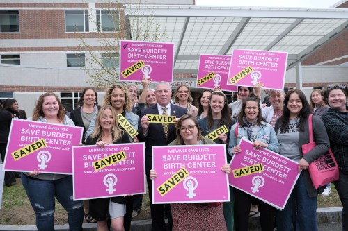 A group of women and one man stand holding signs that say "Save Burdett Birth Center" with a yellow "SAVED" sticker added to the front.