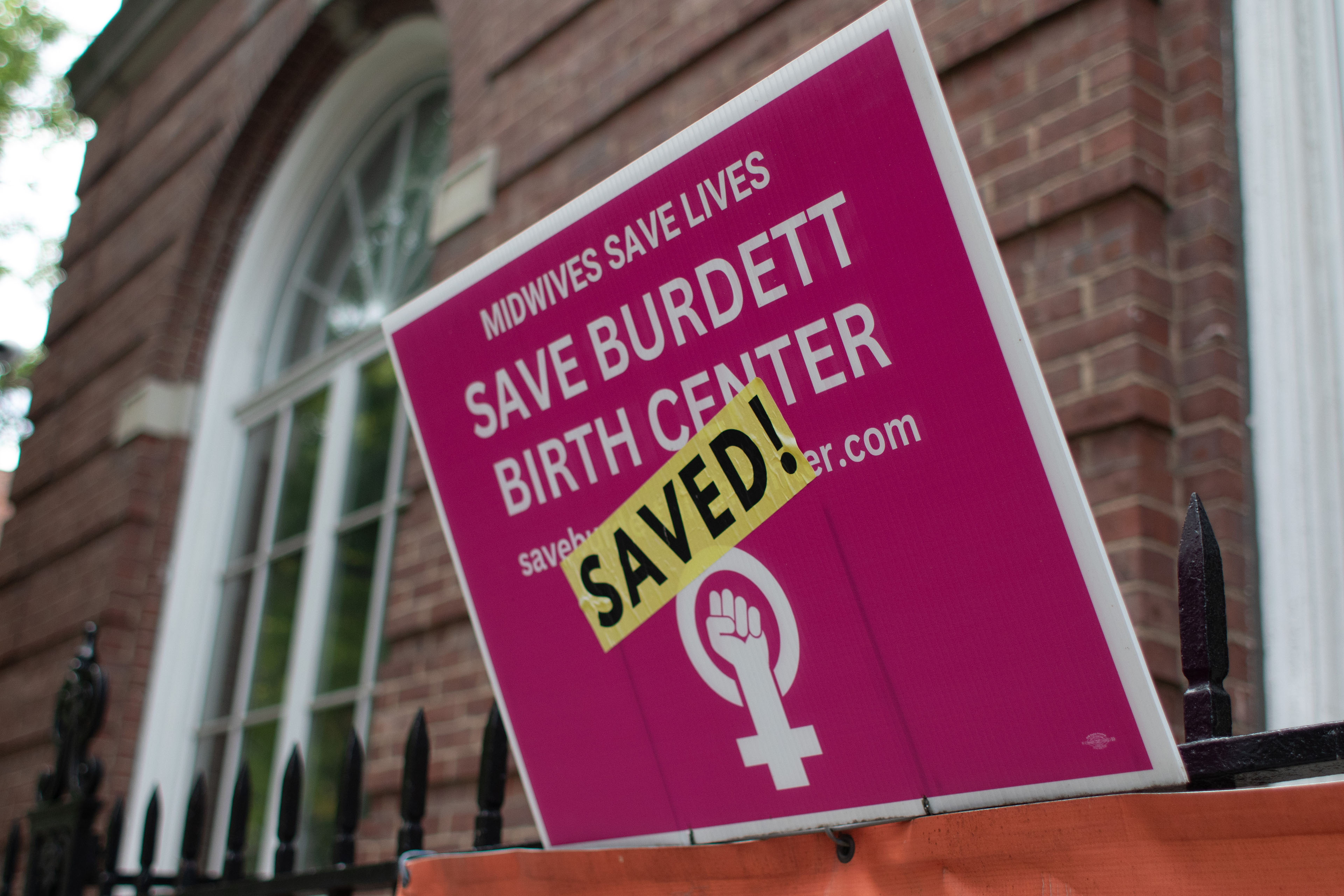 A plastic lawn sign with white text and a bright pink background reads, "Midwives Save Lives / Save Burdett Birth Center / SAVED!"