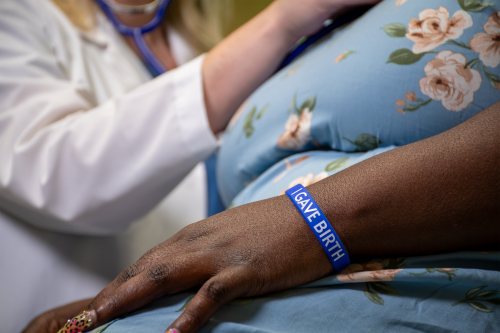 A close-up image of a postpartum woman in a blue dress with flowers. Her arm in the foreground shows a blue wristband that reads, "I gave birth."
