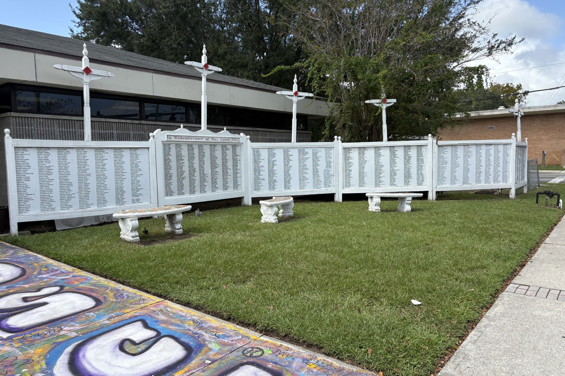 A photo shows a length of white wall on a square of grass. Crosses line the top of the wall. Names are written on the wall.