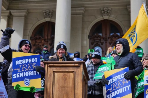A woman speaks at a podium on the steps of a state building. Supporters surround her, some holding signs that say, "Respect RN seniority."