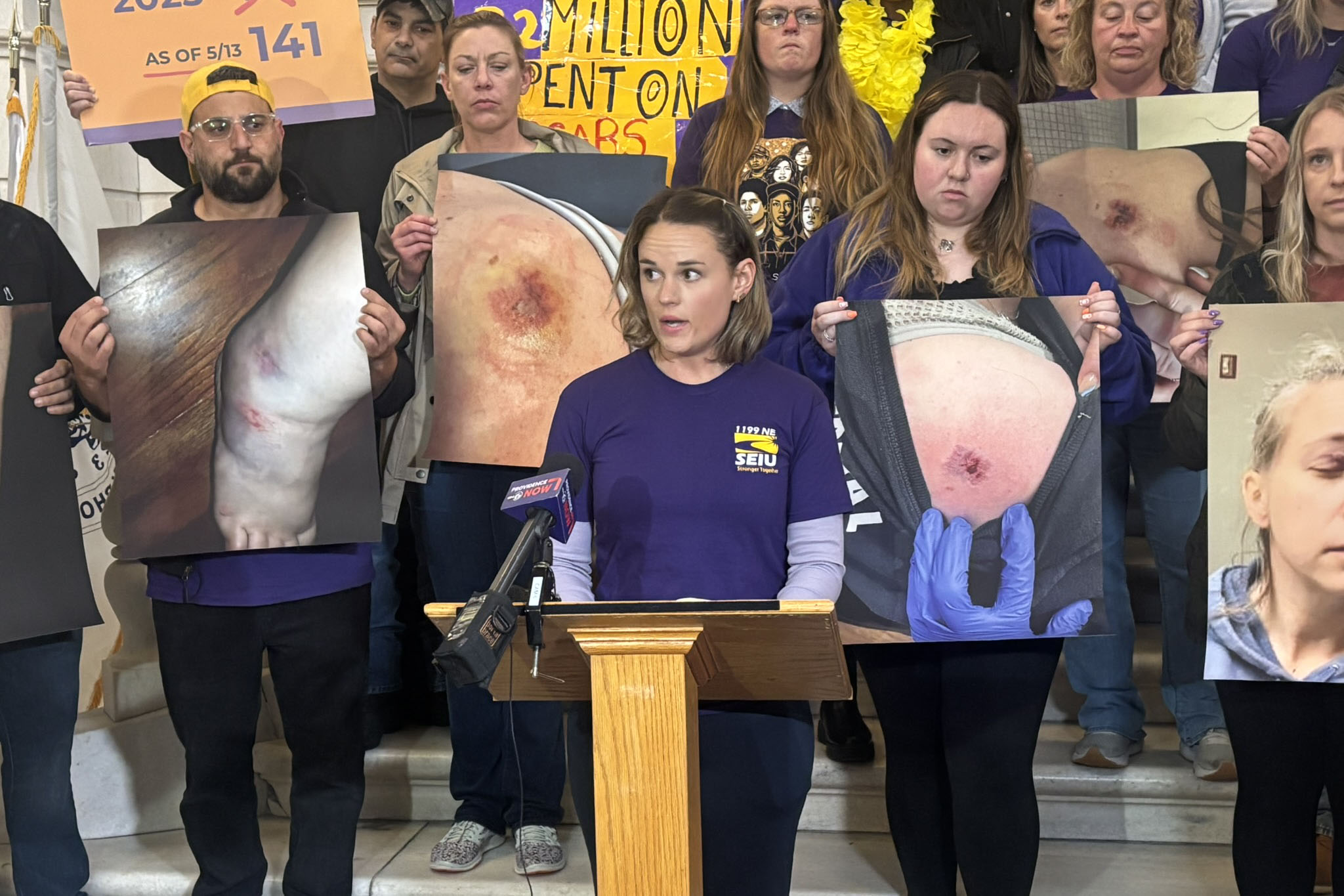 A woman wearing a purple T-shirt speaks at a podium on the steps of a state building. Behind her, supporters hold large pictures of injuries they've suffered on the job.