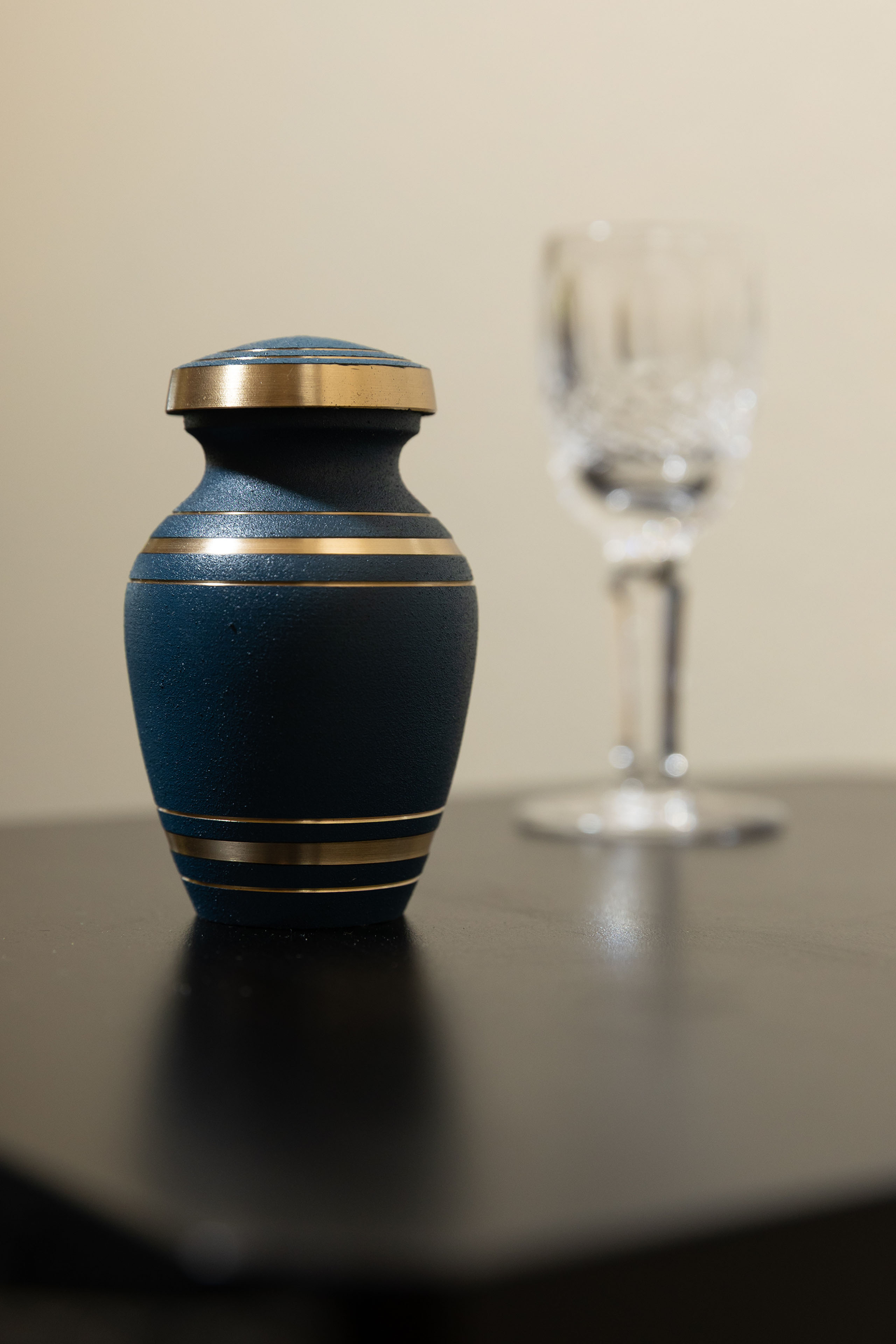 A photo of a dark blue urn on a black table. A glass goblet is seen behind the urn.