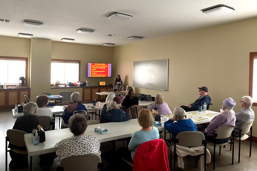 A group of people sit at tables facing a speaker at the front of the room in an educational setting.