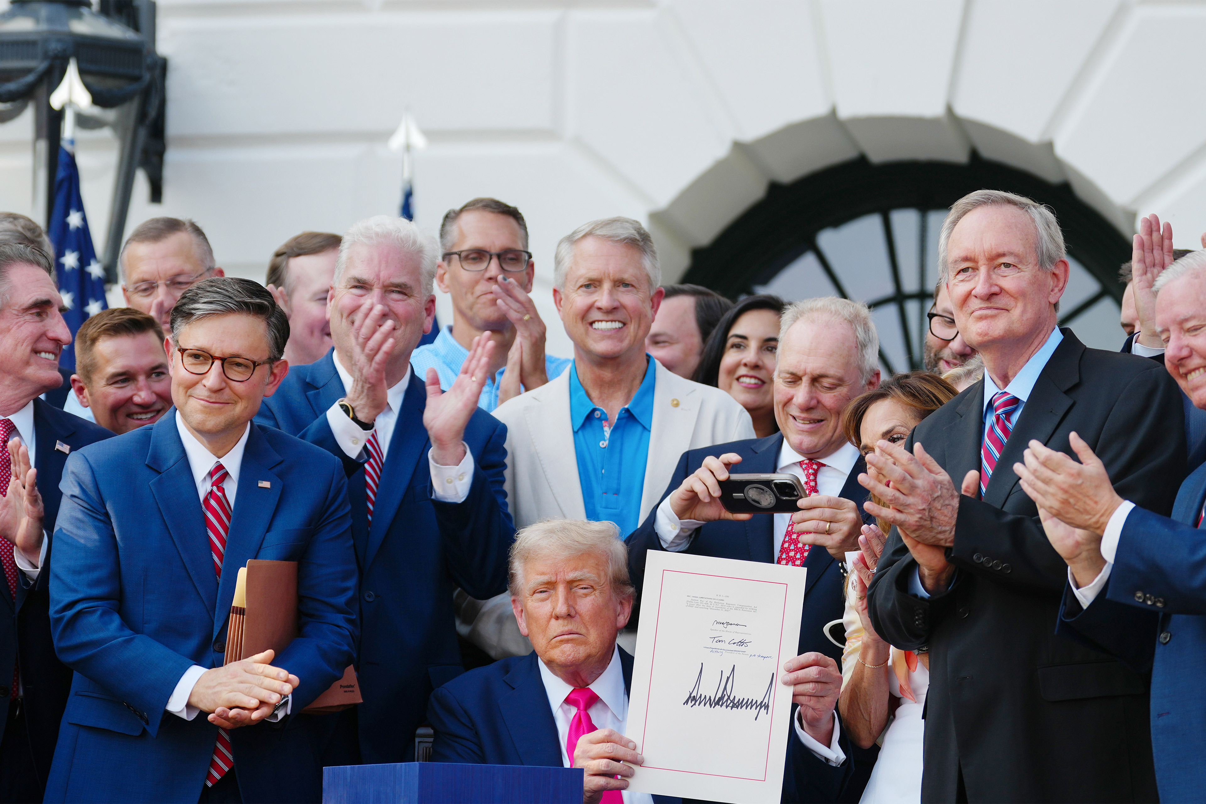 A photo of President Trump surrounded by various Republican members of Congress at an event on the South Lawn of the White House. Trump, seated, holds up a signed bill.