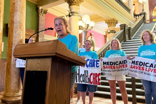 A photo of Rep. Mary Caferro speaking at a podium. Behind her stand several women holding signs that read, "Keep the care in healthcare," and "Medicaid saves lives. Hands off Medicaid."