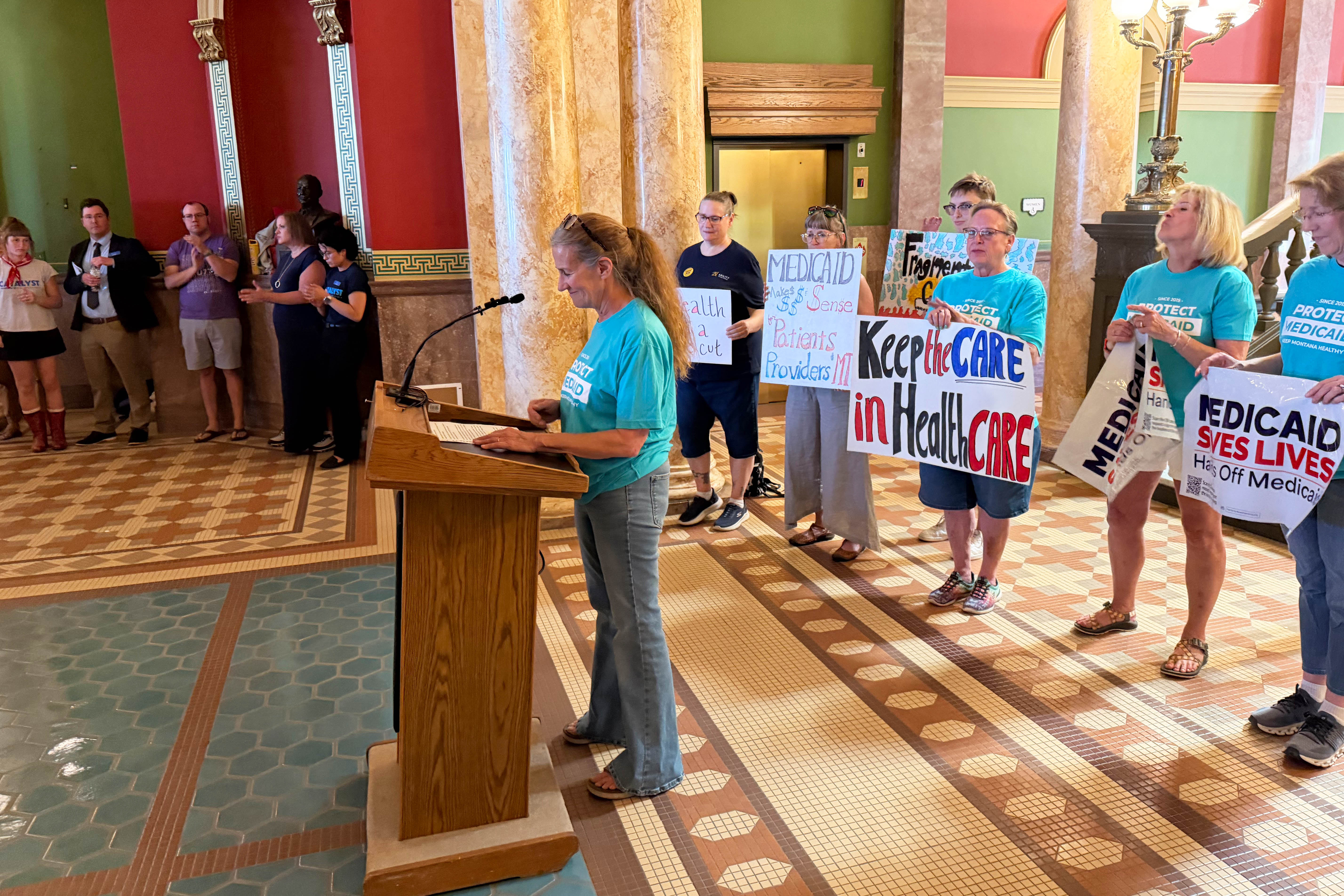 A photo of Rep. Mary Caferro speaking at a podium seen from a different angle. Behind her stand several women holding signs that read, "Keep the care in healthcare," and "Medicaid saves lives. Hands off Medicaid."