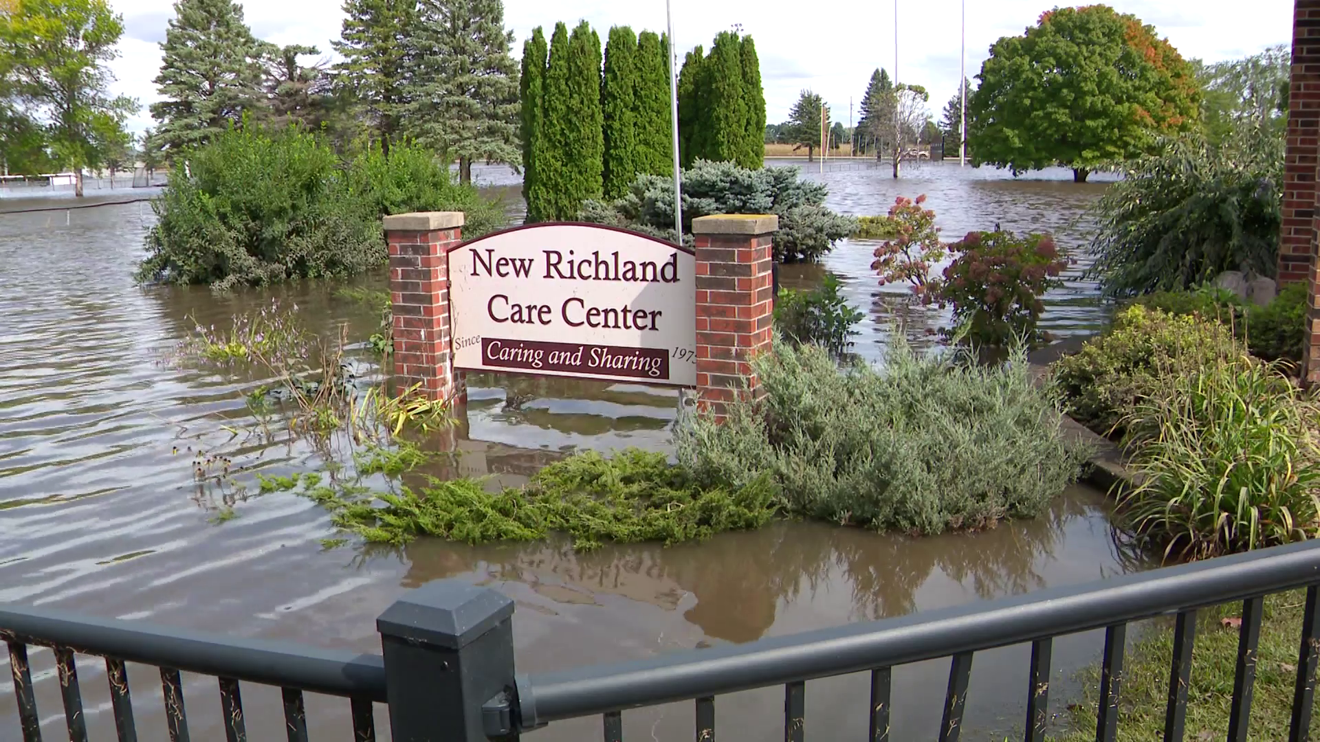 The area in front of a building is flooded and a freestanding sign reads "New Richland Care Center, Caring and Sharing, Since 1973."