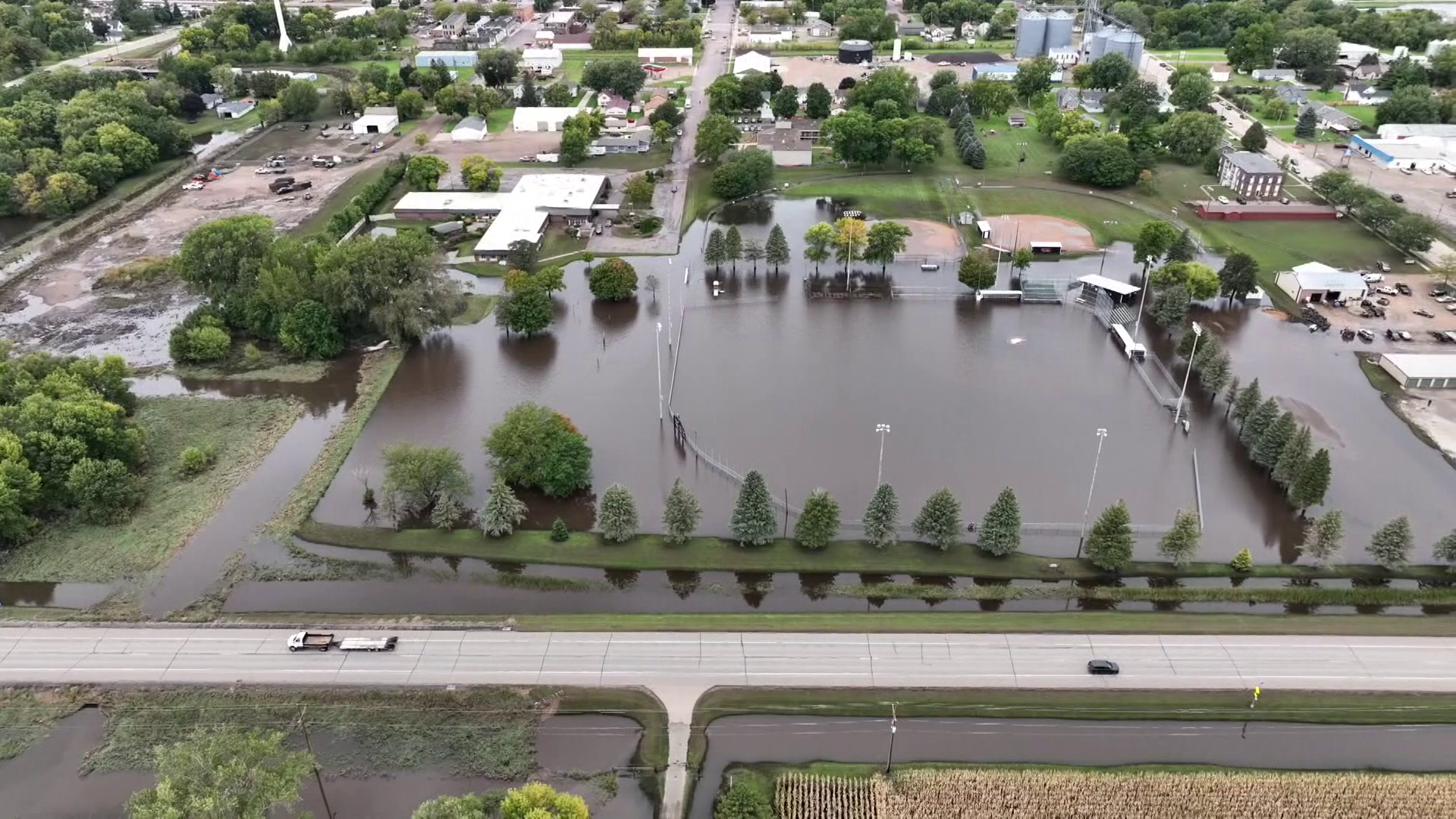 In an aerial image, a baseball field beside a road is flooded with water. The road is dry.