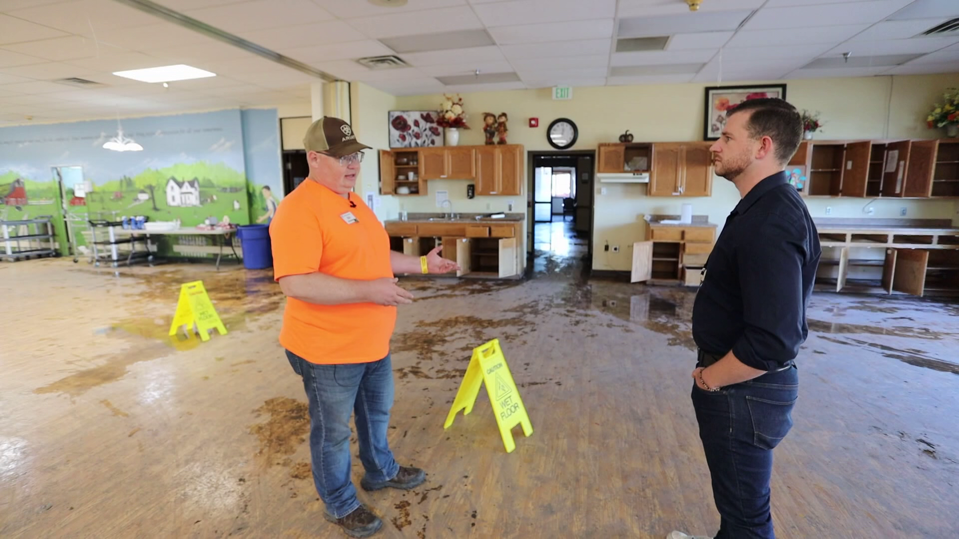 Two people stand inside a large room and talk. Areas of the floor have standing water and two "Caution: Wet Floor" signs have been placed.