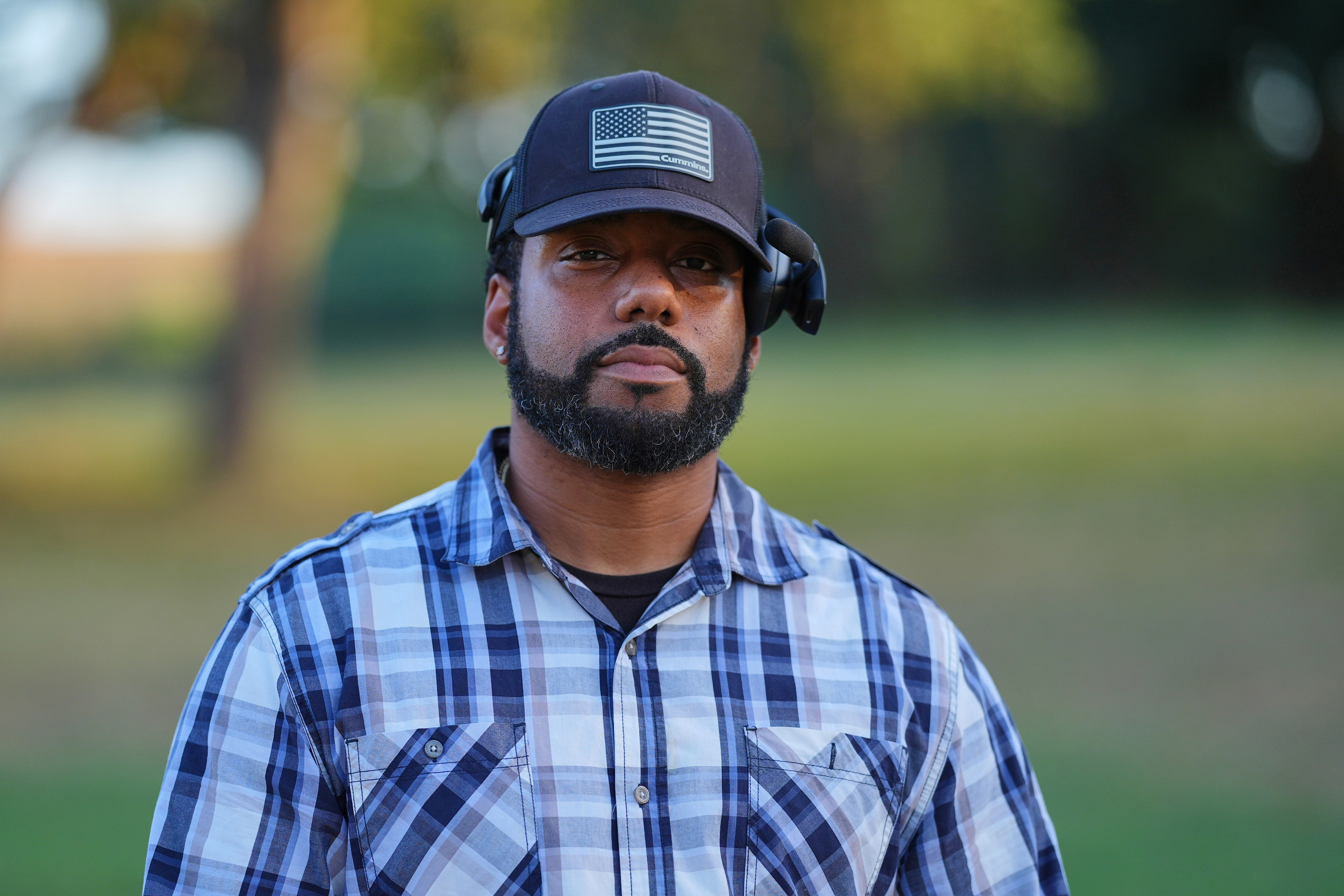 A man wearing a plaid shirt and a black hat with an American flag poses for a photo