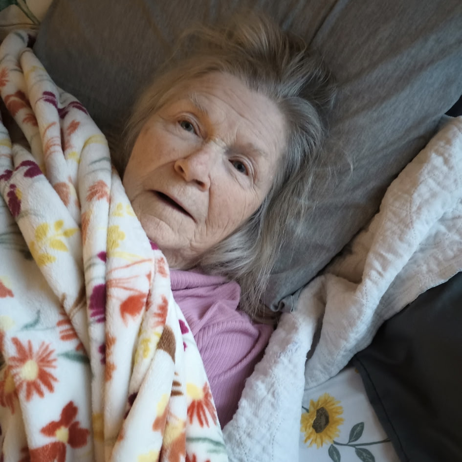 A senior woman is lying on her back in bed and looking up at the camera.