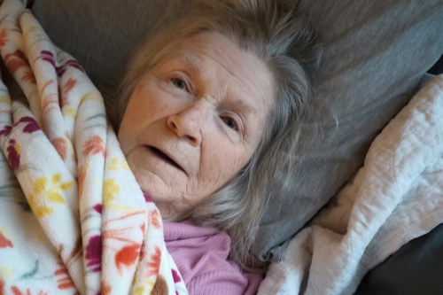 A senior woman is lying on her back in bed and looking up at the camera.