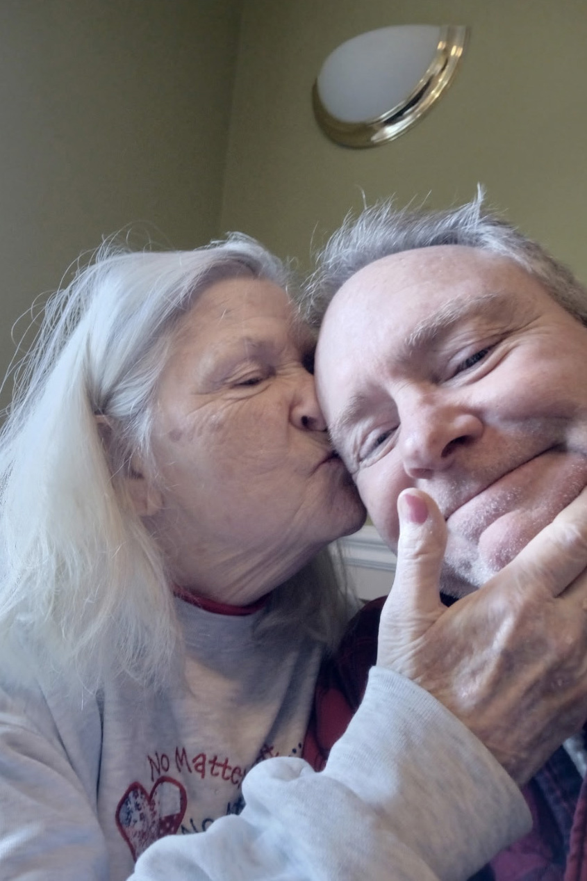 A senior woman with long white hair lovingly kisses her adult son on the side of his face.