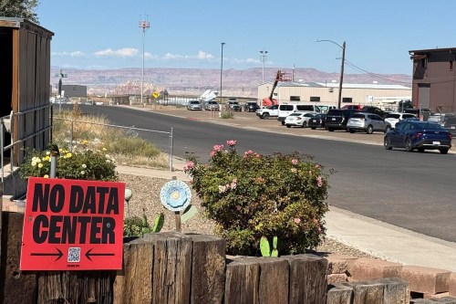 A photo of a street in Page, Arizona. In the foreground is a small red sign with a QR code that reads, "No data center," staked into the yard of a house. In the background are cars parked in lots around Page Municipal Airport.