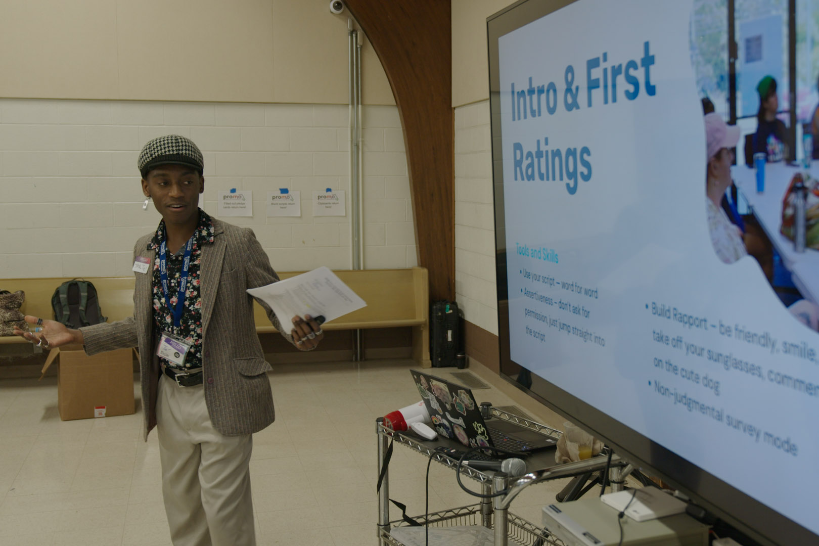 Te’ Hopkins gives a presentation in front of a large screen. The screen reads, "Intro & First Ratings. Tools and skills: Use your script — word for word. Awareness — don't ask for permission, just jump straight into the script. Build Rapport — be friendly, smile, take off your sunglasses, comment on the cute dog. Non-judgmental survey mode."