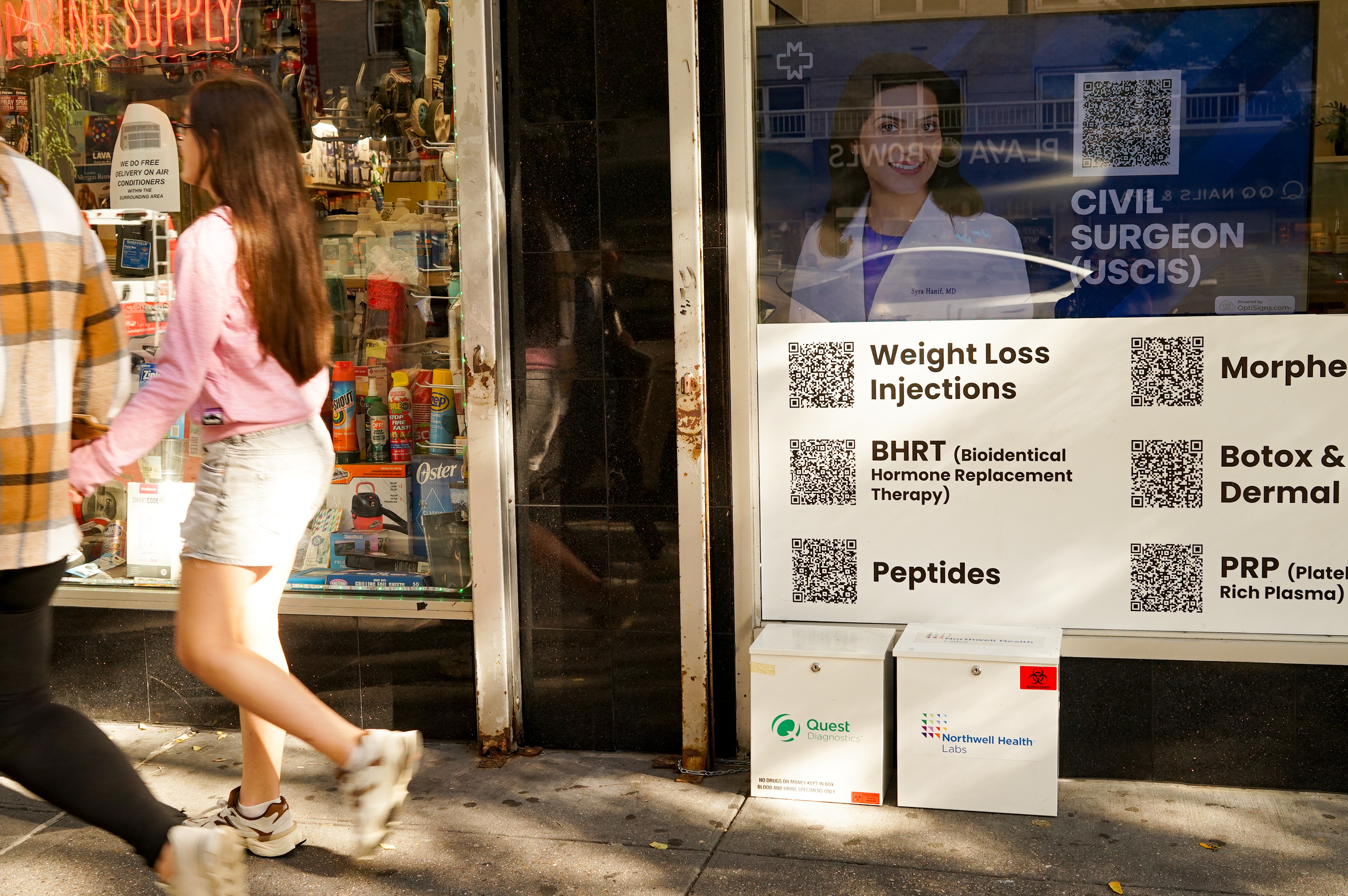Two passersby walk past a wellness clinic. It has a sign that advertises peptides alongside weight-loss injections, bioidentical hormone replacement therapy, and botox.