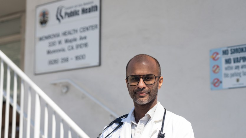A doctor in a white coat with a stethoscope stands in front of a staircase and a sign that reads "Monrovia Health Center"