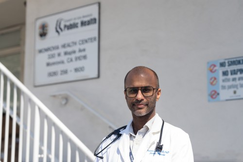 A doctor in a white coat with a stethoscope stands in front of a staircase and a sign that reads "Monrovia Health Center"