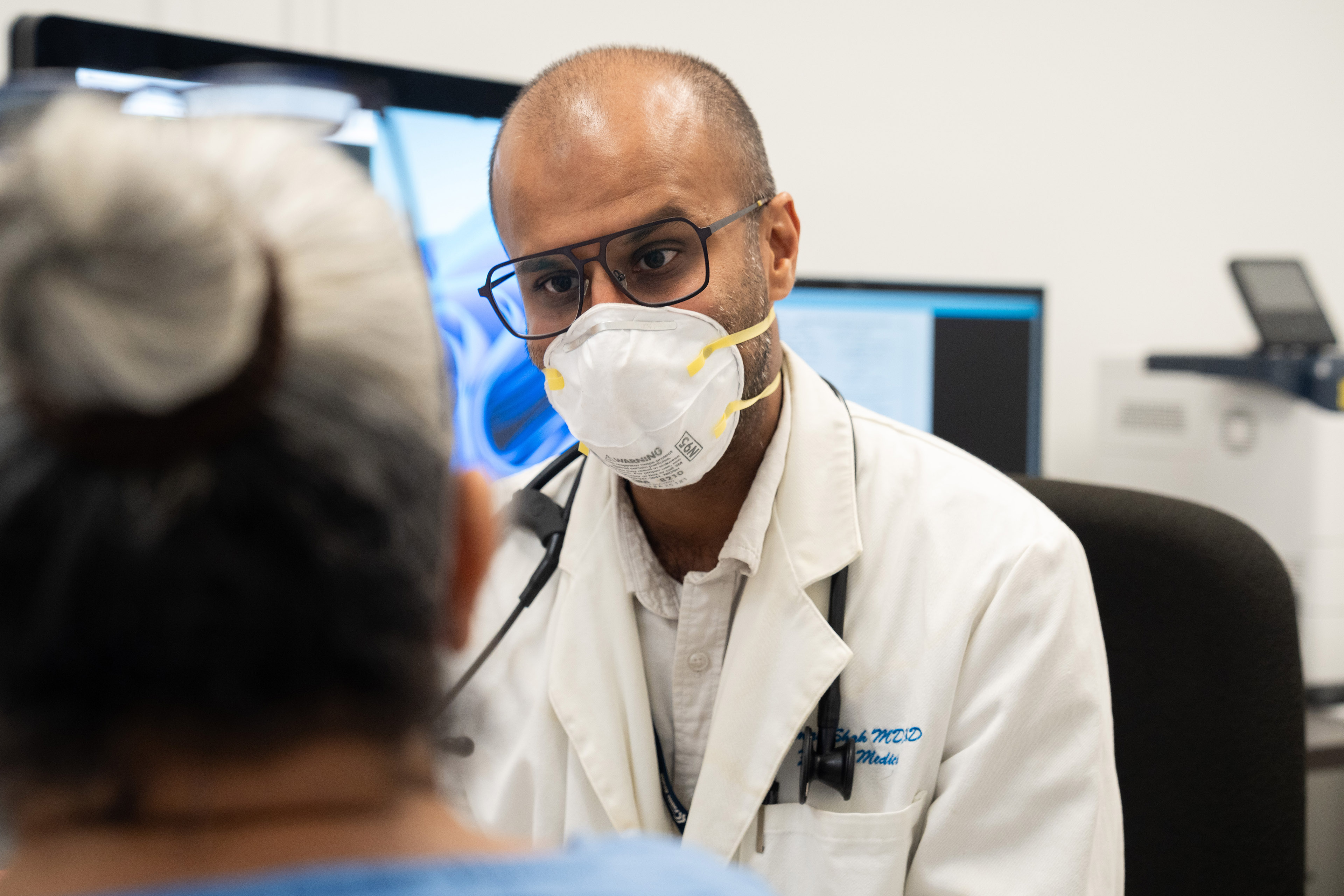 A doctor wearing glasses and a facemask talks with a patient