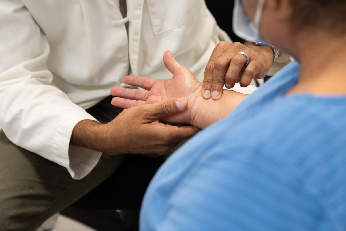 A doctor sitting down, wearing a white coat, feels the wrist of a patient