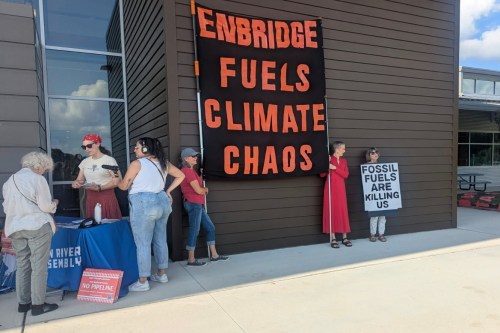 A group of people stand outside in protest. Two of them hold up a large sign that reads, "Enbridge Fuels Climate Chaos." Another holds a sign reading, "Fossil fuels are killing us."