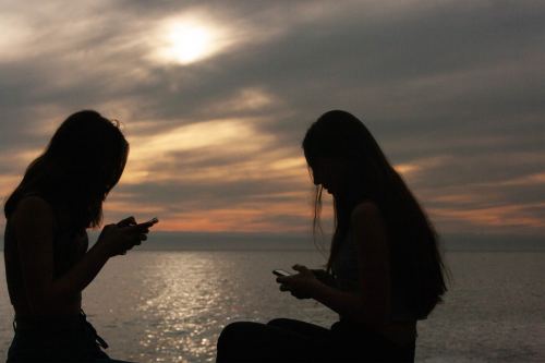 A photo shows silhouettes of two teenage girls using their phones at sunset.
