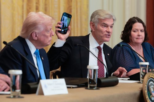 A photo of President Donald Trump, Mehmet Oz — holding up a cell phone — and Amy Gleason seated at an event in the White House.