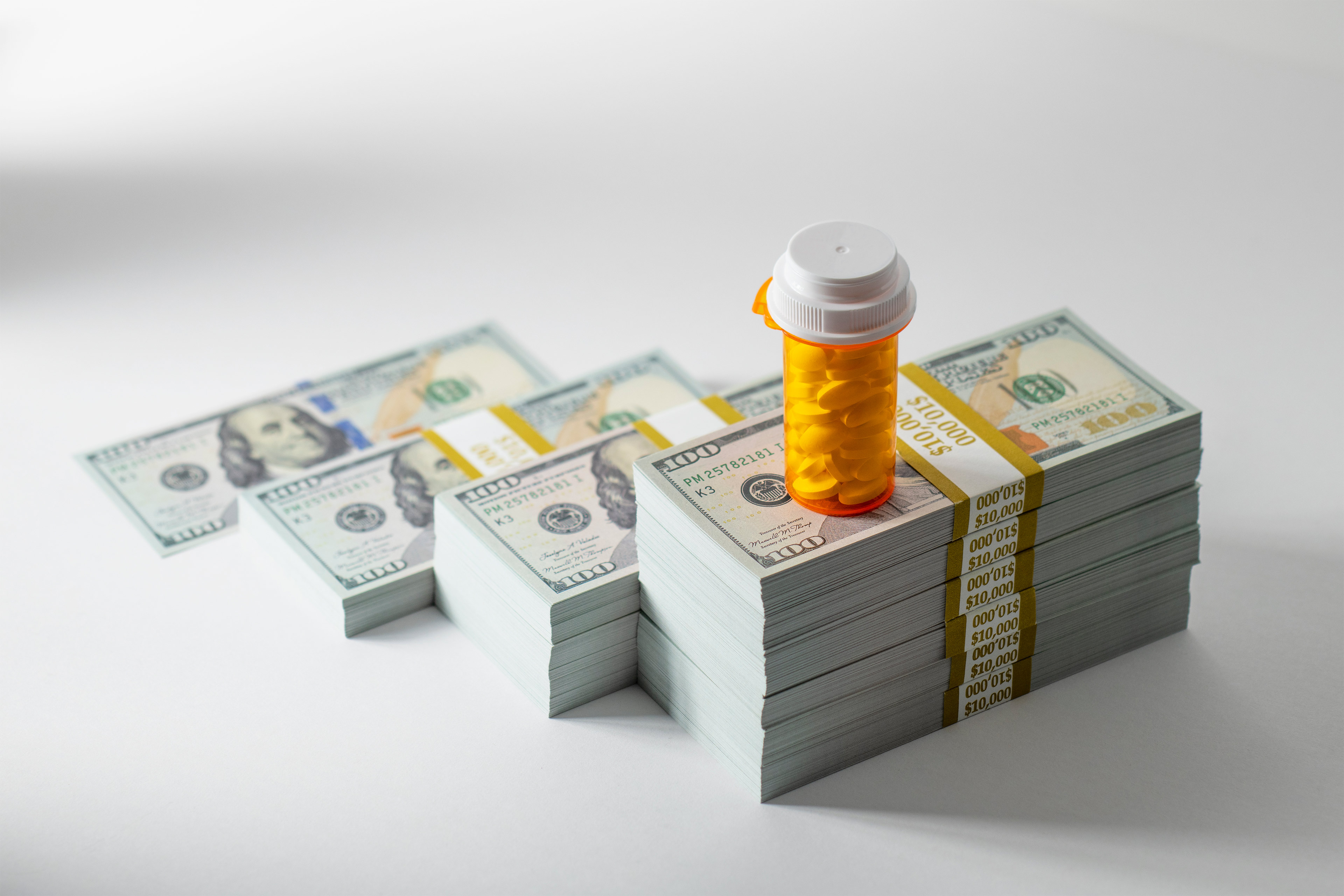 A photo shows four increasingly-taller stacks on one-hundred-dollar bills. The tallest stack on the right has an orange pill bottle resting on top of it.