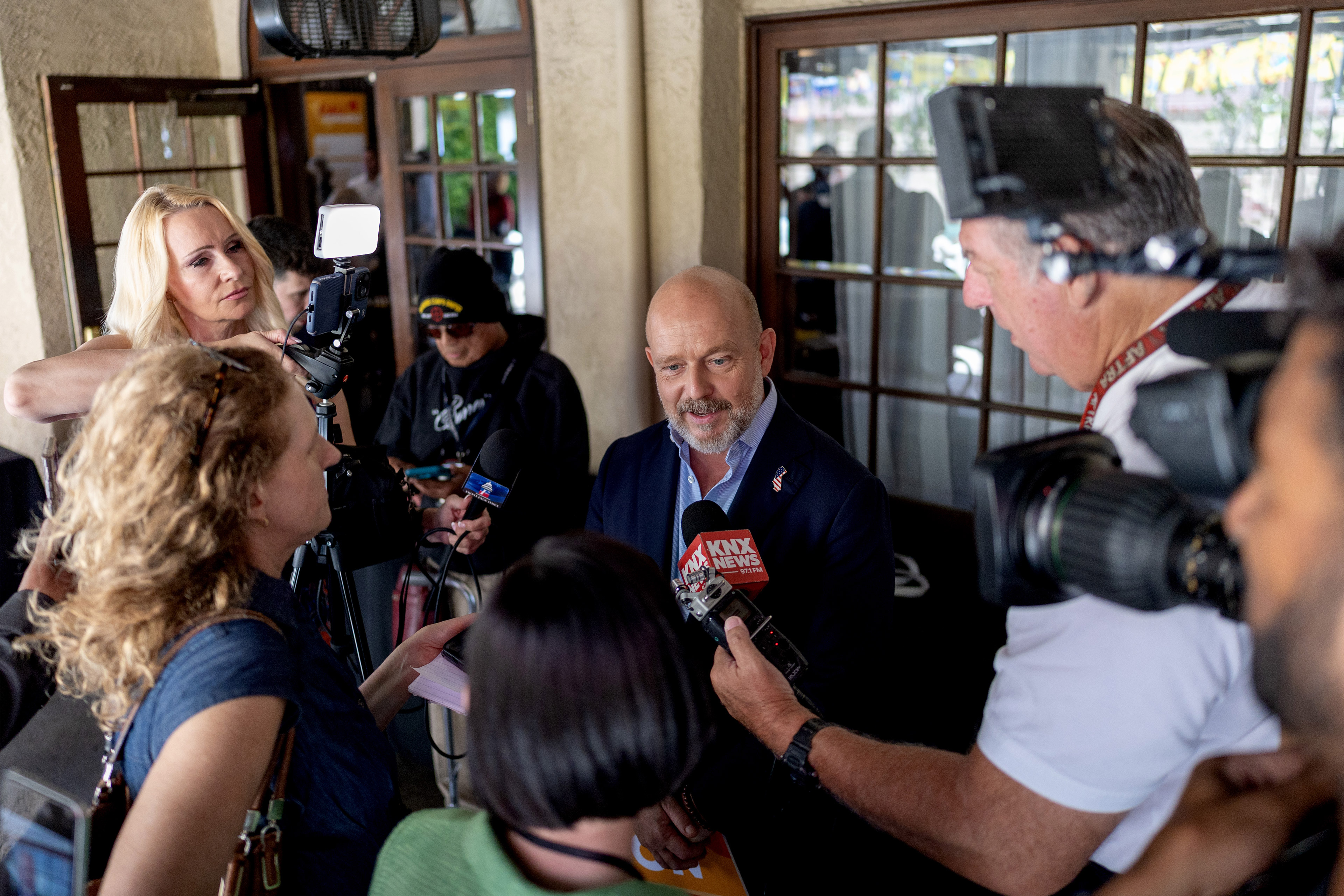Steve Hilton is surrounded by reporters for various media outlets holding out microphones.