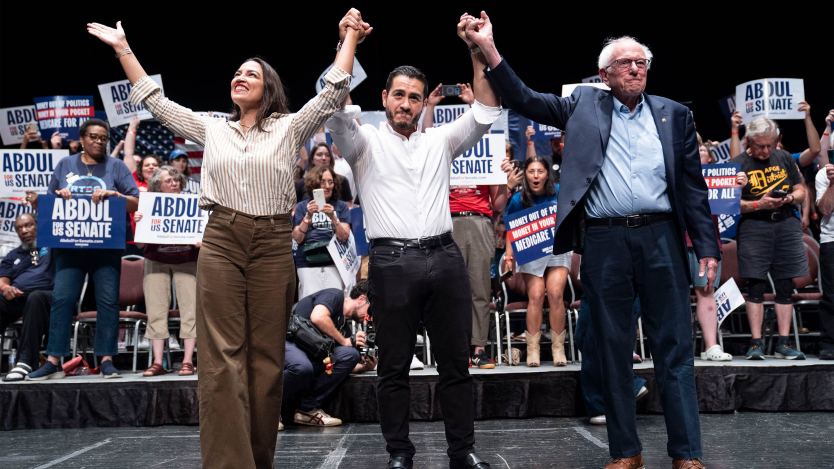 A photo of Alexandria Ocasio-Cortez, Abdul El-Sayed, and Bernie Sanders on stage at a rally. The three are holding hands with their arms raised.