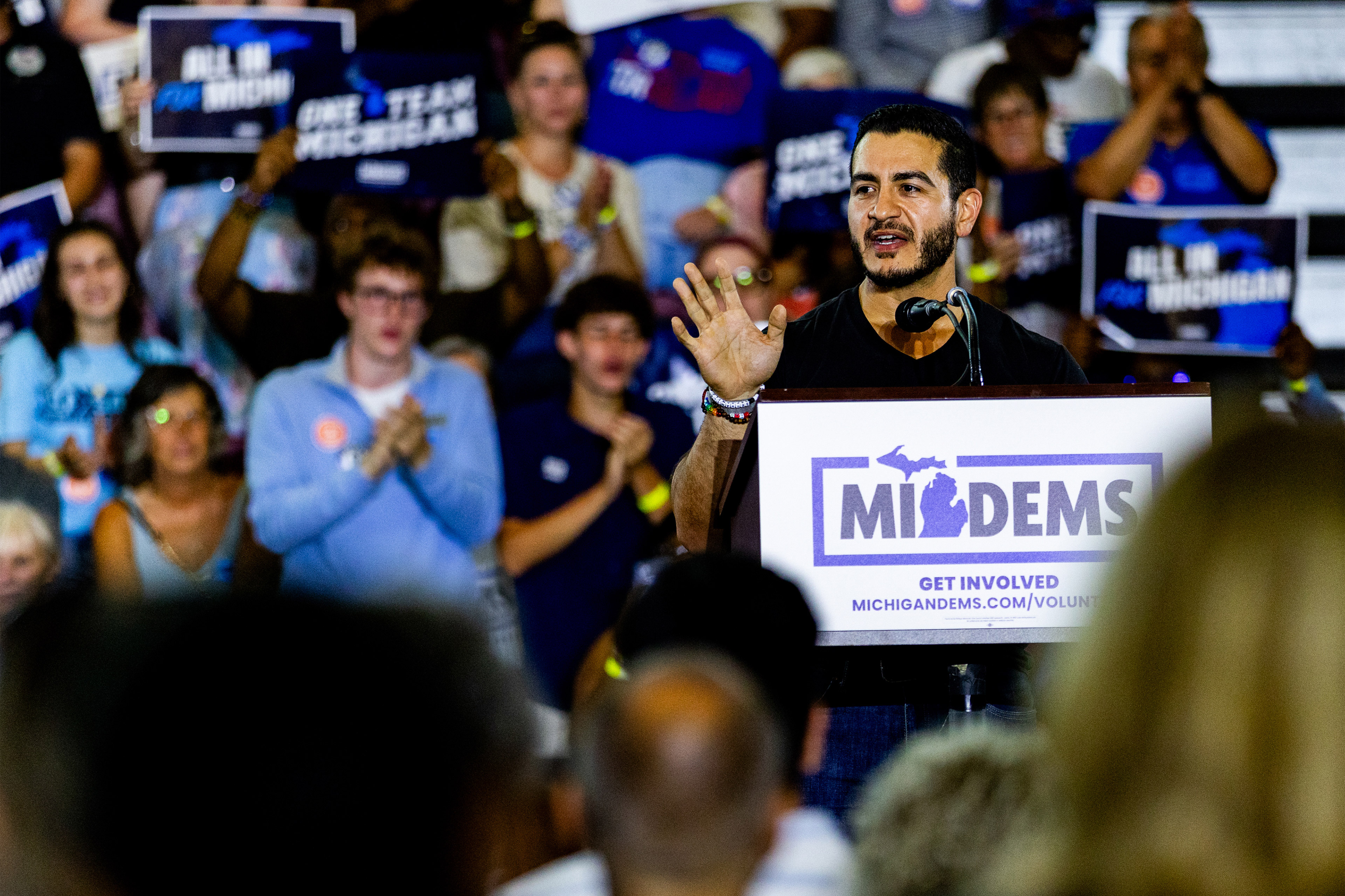 Abdul El-Sayed stands at a podium in an event space. He is surrounded on both sides by supporters holding signs.
