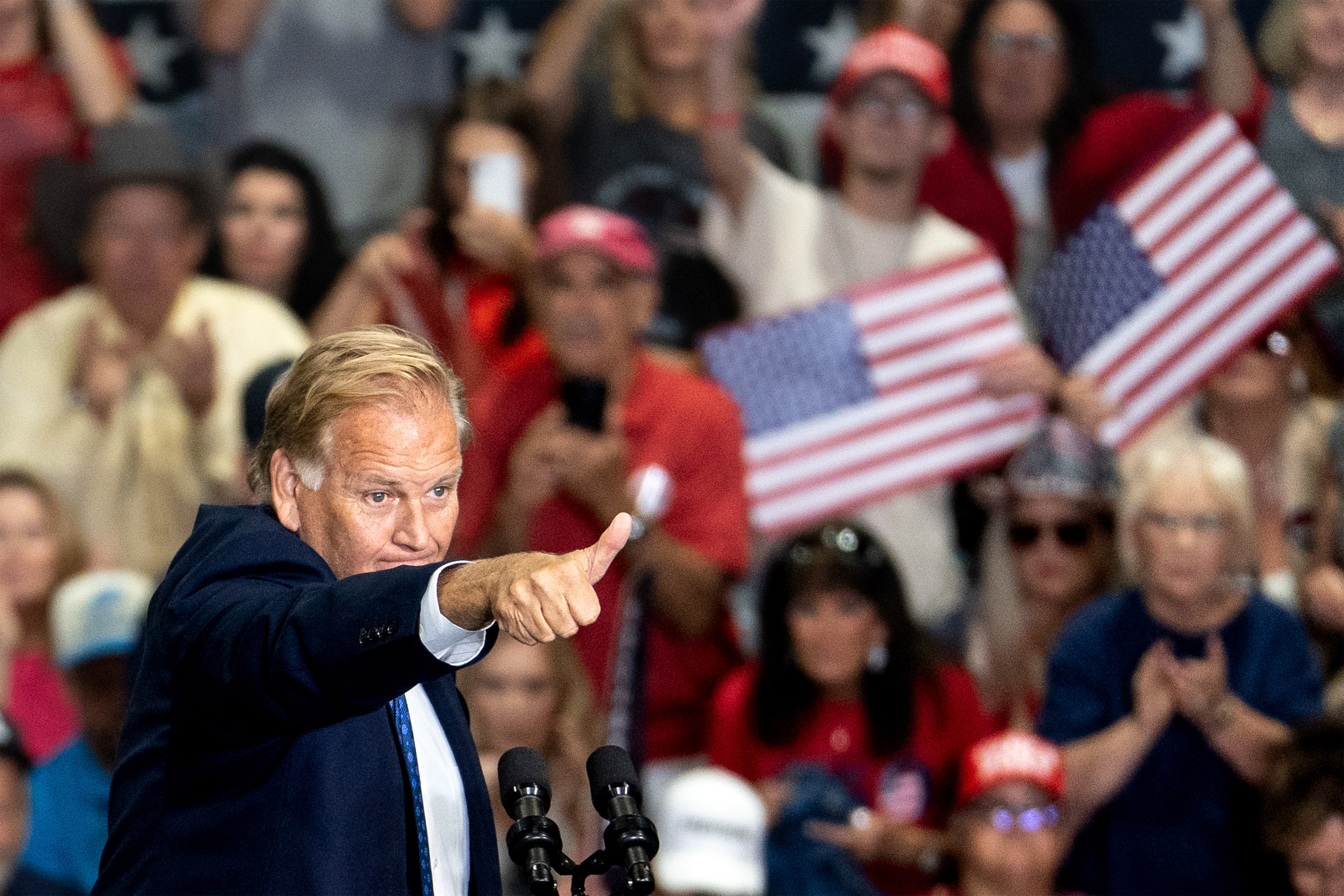 Mike Rogers gives a thumbs up on stage at a rally. Supporters behind him hold American flags.