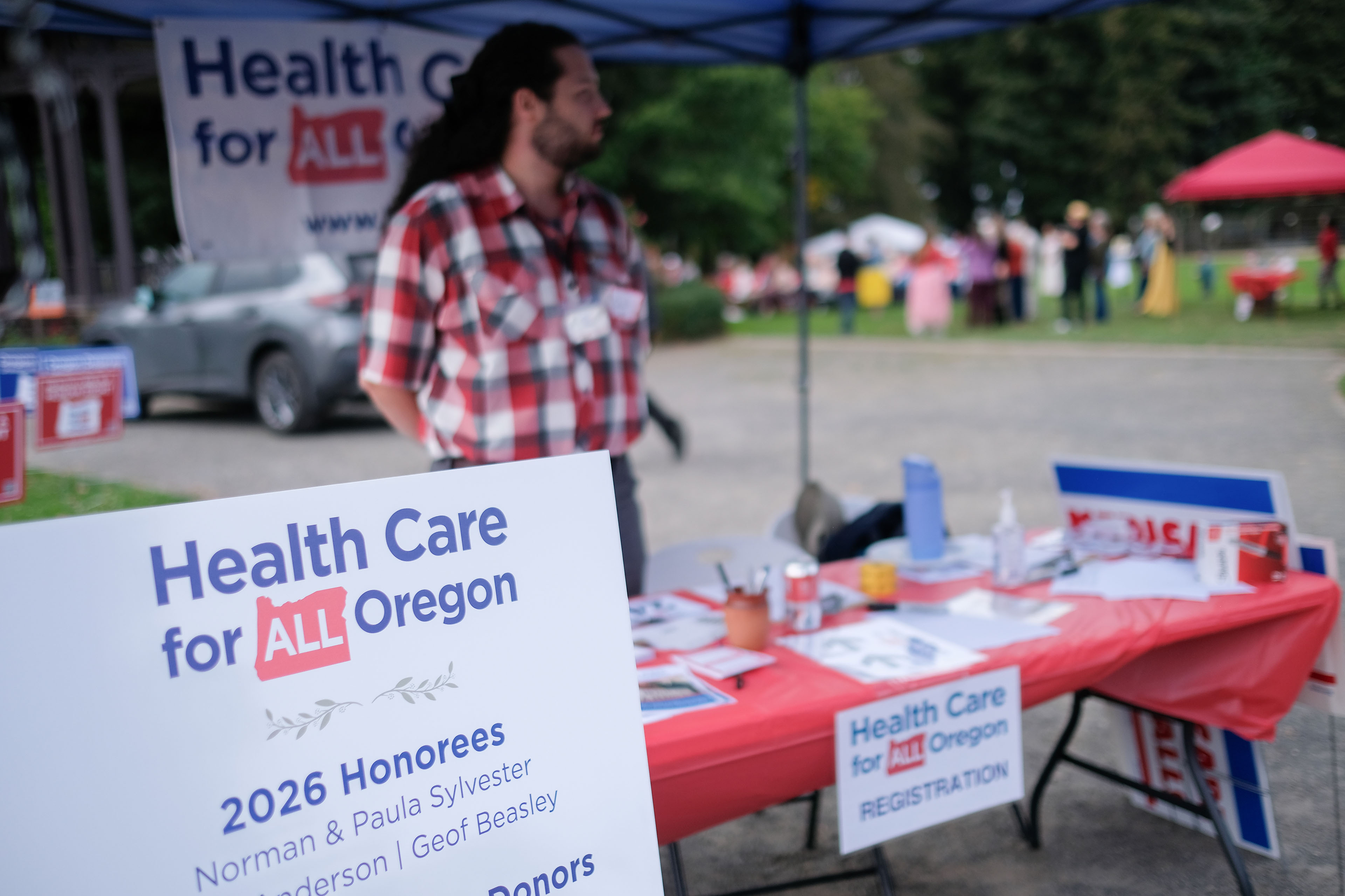A man stands at a vendor table outdoors. He has signs, pins, and other information about "Health Care for All Oregon."