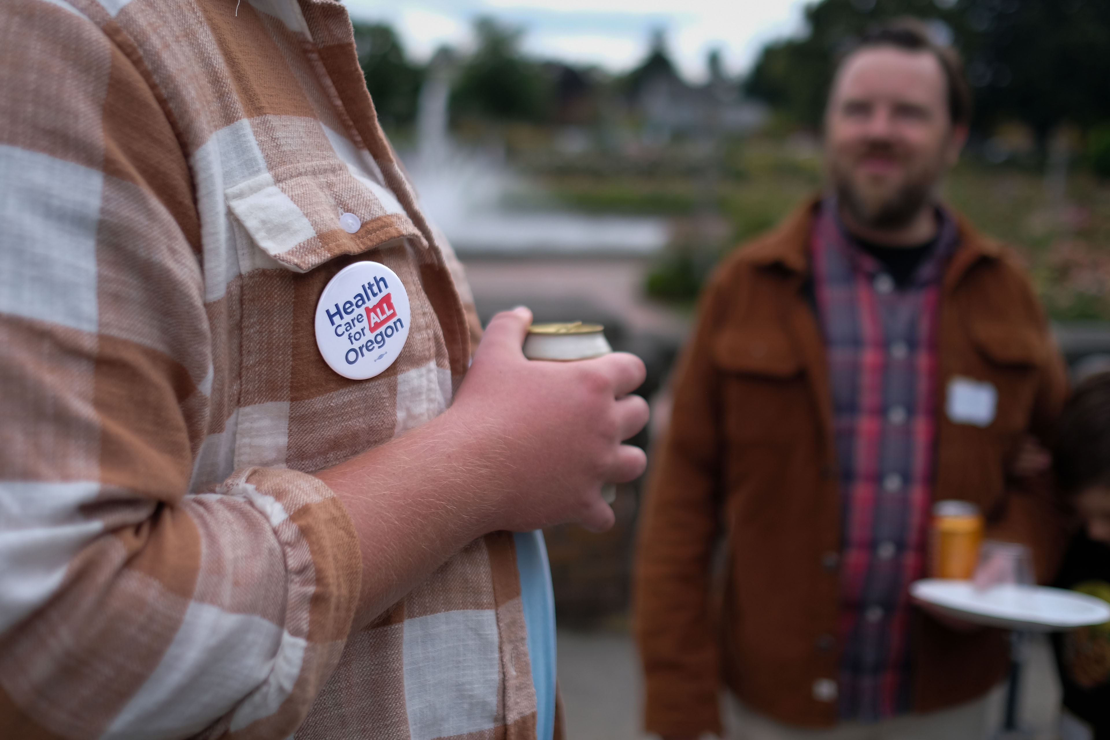A man wearing plaid is holding a canned beverage while speaking to another man close beside him. A button is visible on the man in plaid's shirt, it says "Health Care for All Oregon."