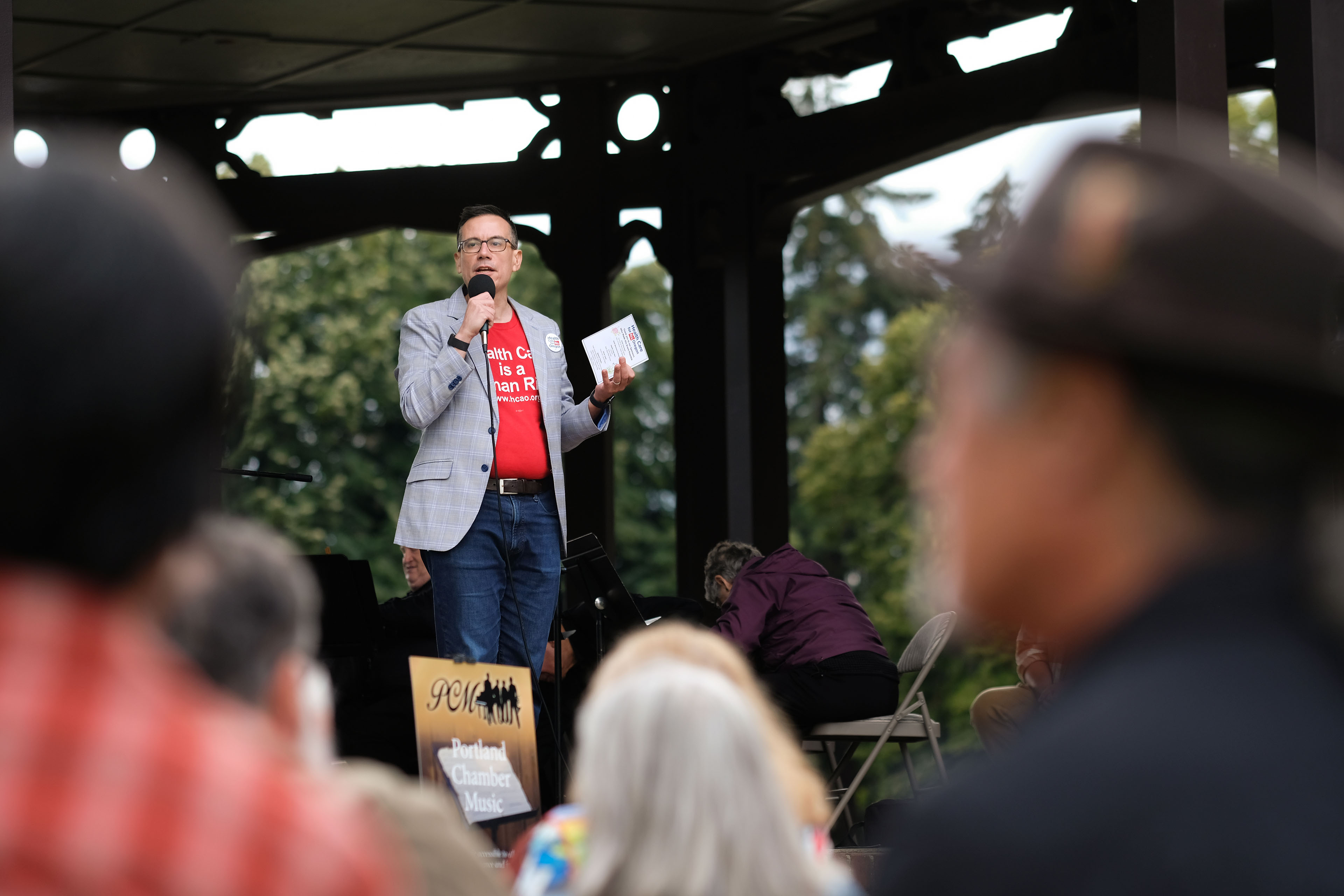 A man wearing a blazer over a bright red shirt stands in front of a crowd while speaking into a microphone in an outdoor setting.